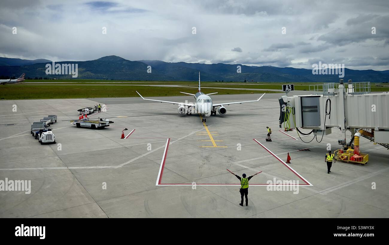 MISSOULA MONTANA AIRPORT, MT, JUN 2022: wide view American Airlines Embraer E175 jet aircraft approaches gate with ground crew and vehicles around it, mountains in background - Smartphone Captured Stock Image