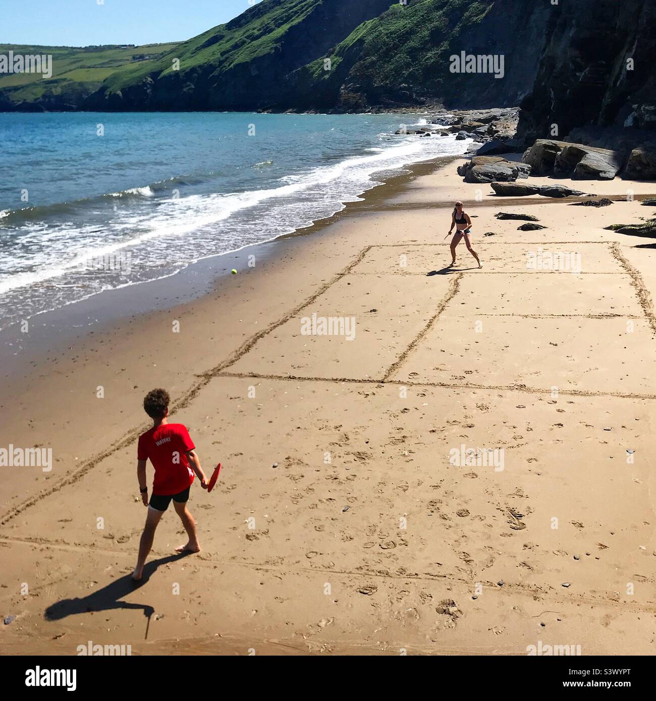 A mother and son playing tennis on a secluded beach near Llangrannog in Ceredigion, Wales. The tide is coming in and the improvised tennis court is hand drawn in the sand. - Smartphone Captured Stock Image