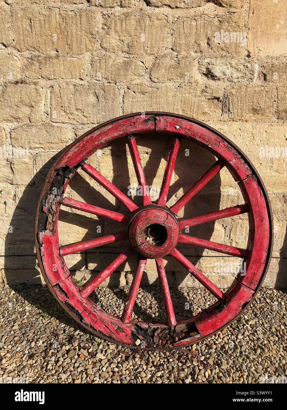 Wooden cartwheel painted red and black with damaged and rotten frame edge leaning against a stone wall - Smartphone Captured Stock Image