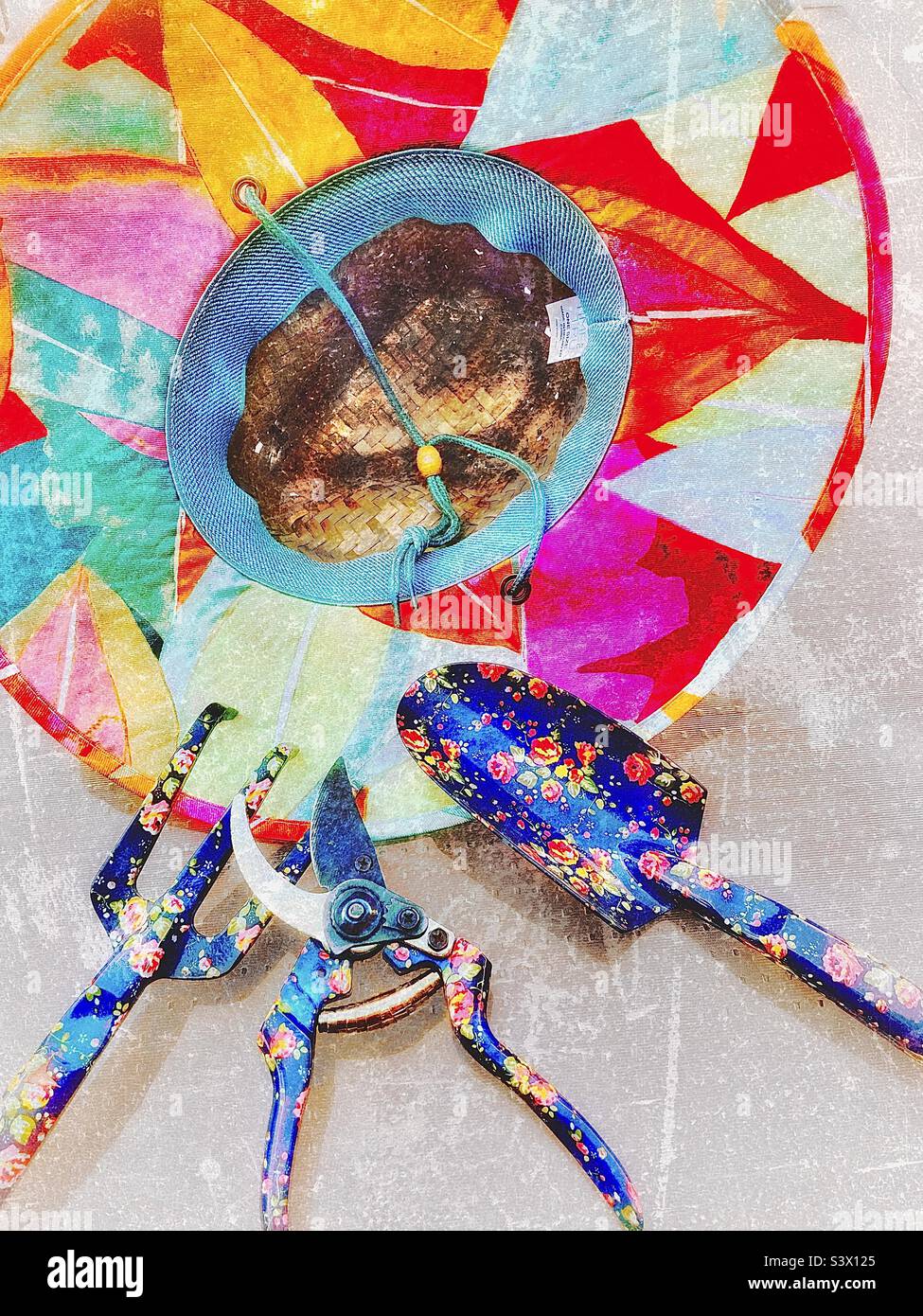 A female gardener’s tools for working out in her yard/garden - a colorful straw hat, and flowery pruning shears, spade and claw. Grunge effect digitally added to this outdoor still life. - Smartphone Captured Stock Image