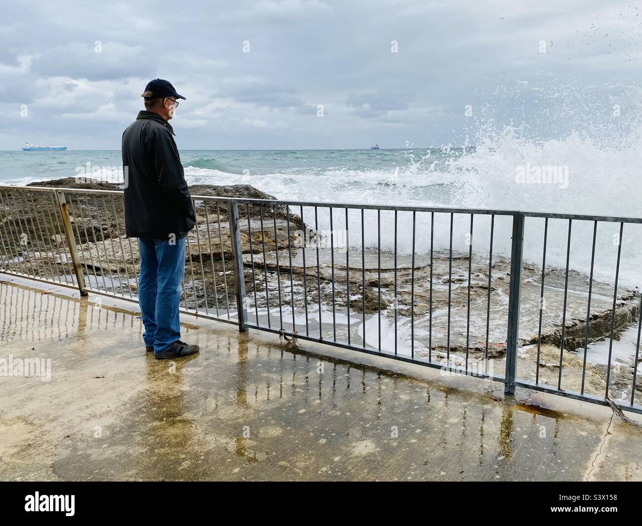Man standing by railings watching the waves hitting a rocky shore Stock Photo