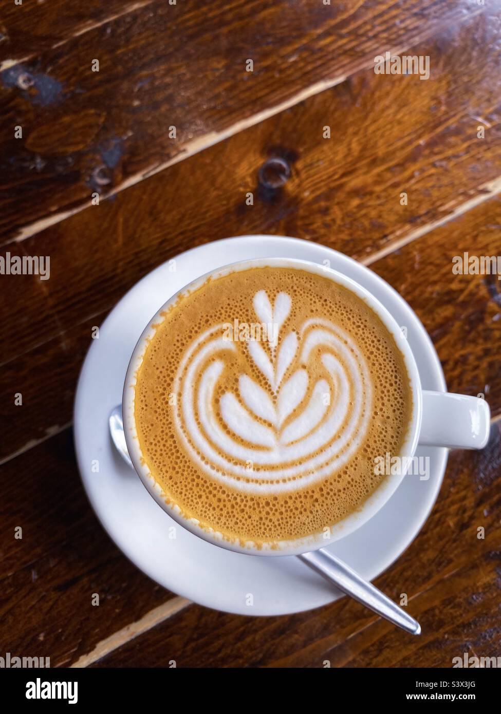 White cup of hot flat white coffee with decorative frothy swirl hearts, sitting on a rustic wooden table. Stock Photo