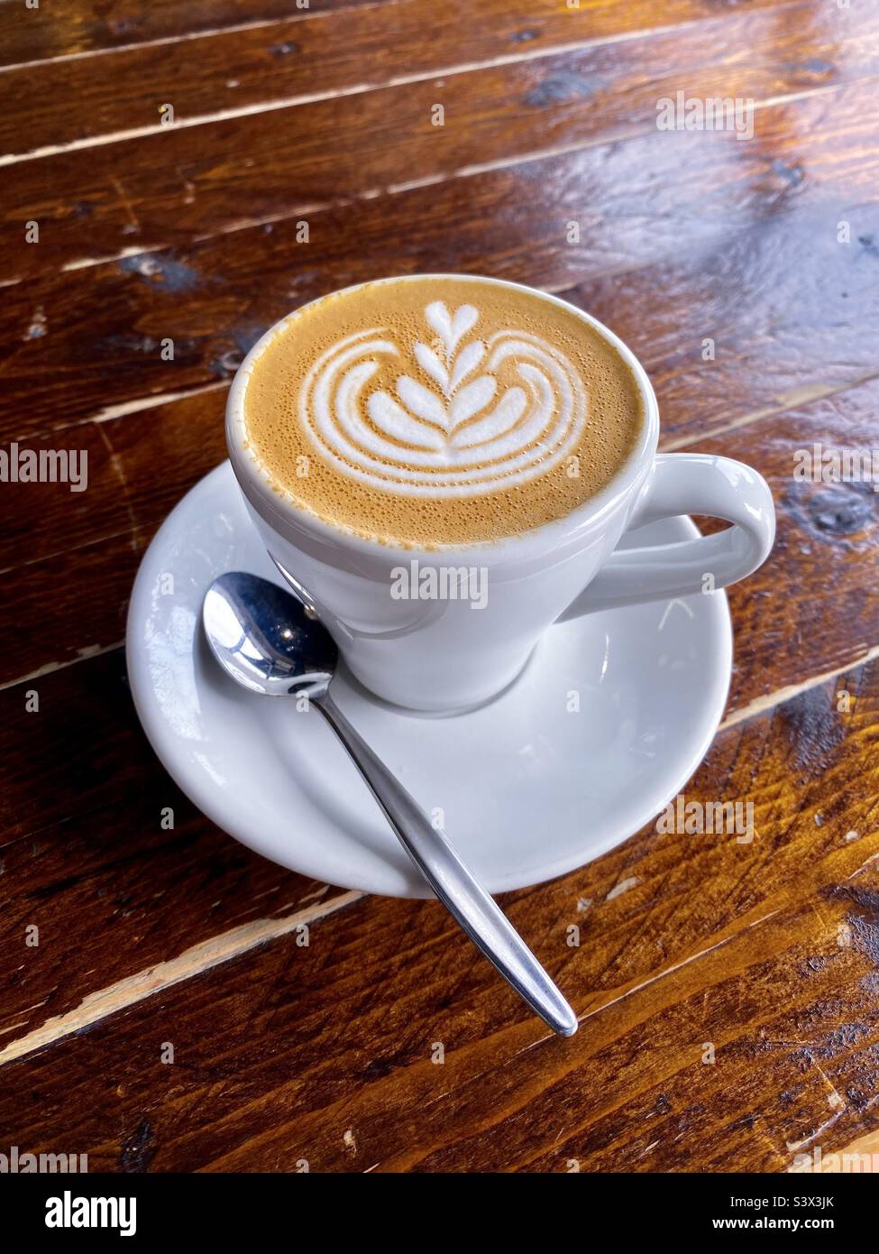 White cup of hot flat white coffee with decorative frothy swirl hearts, sitting on a rustic wooden table. Stock Photo