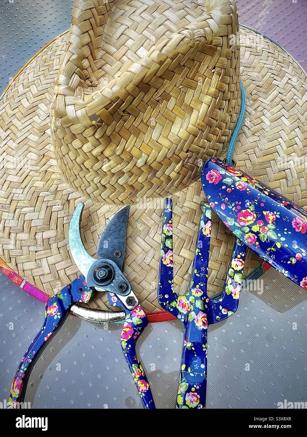A gardening still life made up simply of a female gardener’s straw hat, and her flowery garden tools, pruning shears, a garden claw and a spade or trowel. Nice summertime image. - Smartphone Captured Stock Image