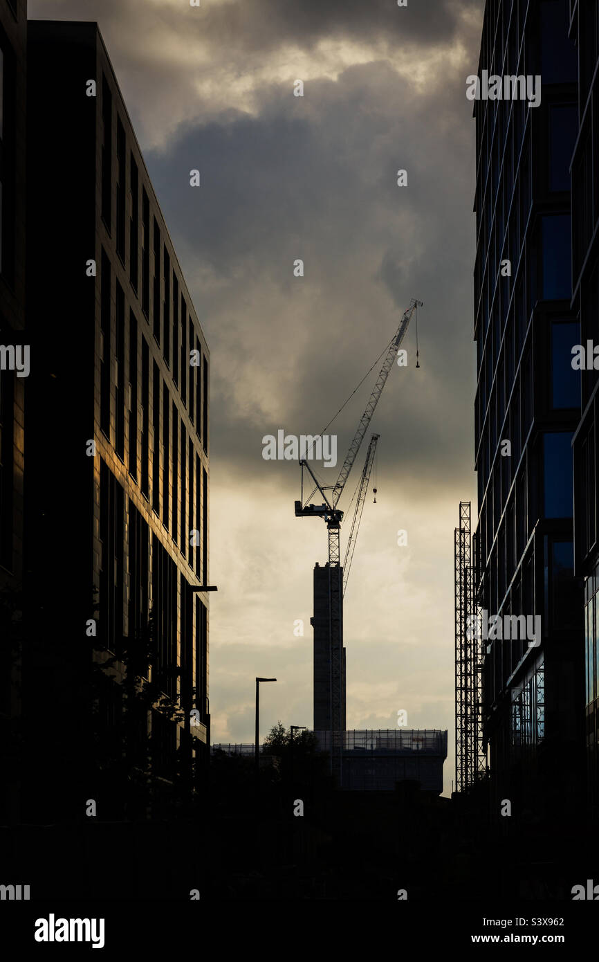 A tall crane building skyscrapers on a construction site between torn city buildings at sunset Stock Photo