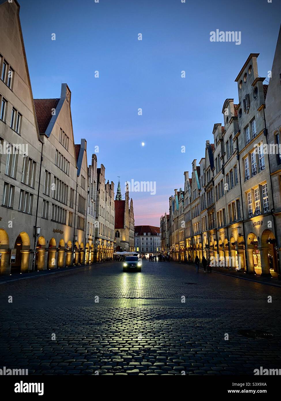A lone car drives down a twilight cobbled street in the old town of the German city of Munster. Darkness on the edge of town. In the moonlight afterglow of the sunset. Like a scene from a spy film. - Smartphone Captured Stock Image