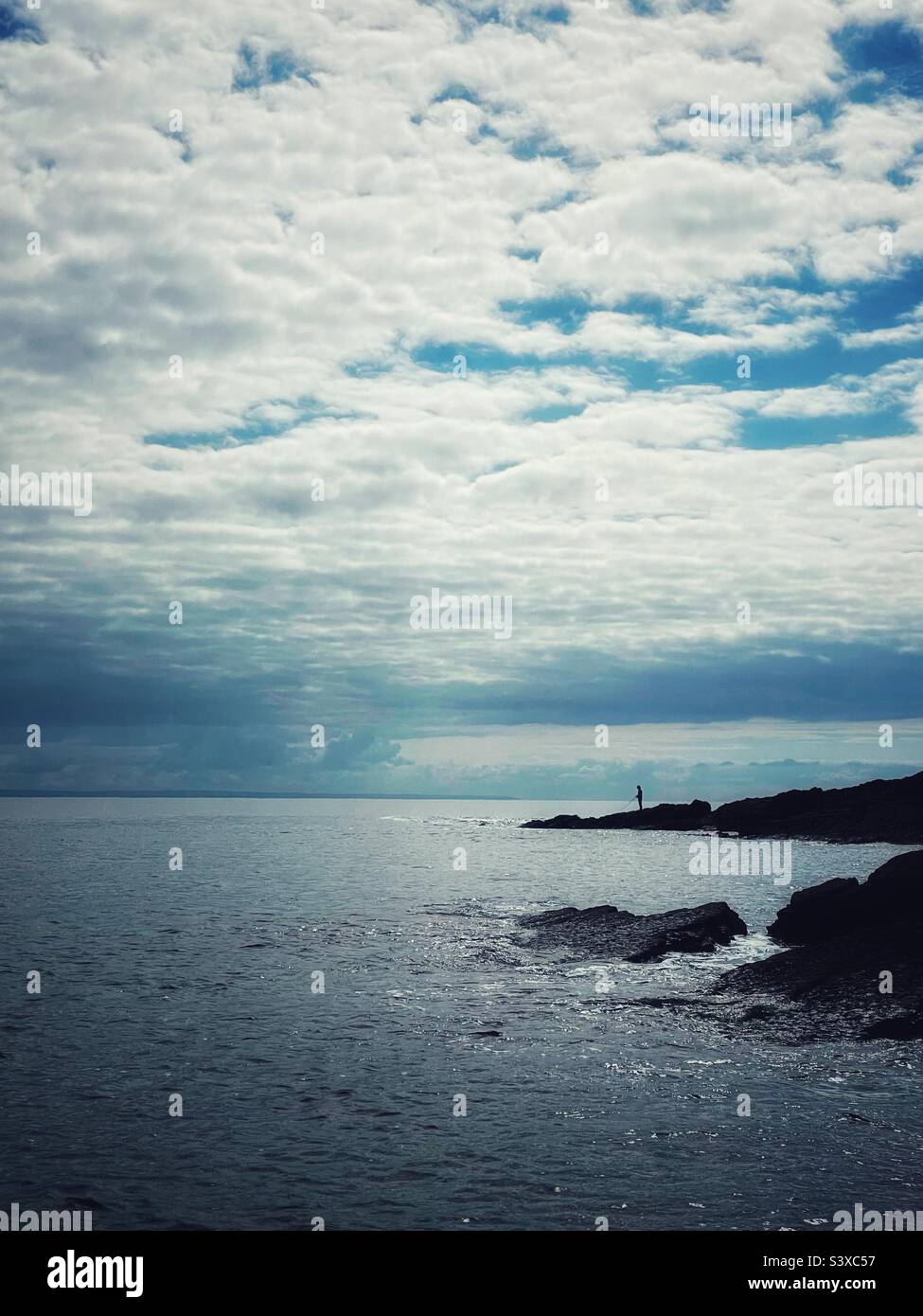 A lone angler fishing from rocks, silhouetted, Wales, September. - Smartphone Captured Stock Image
