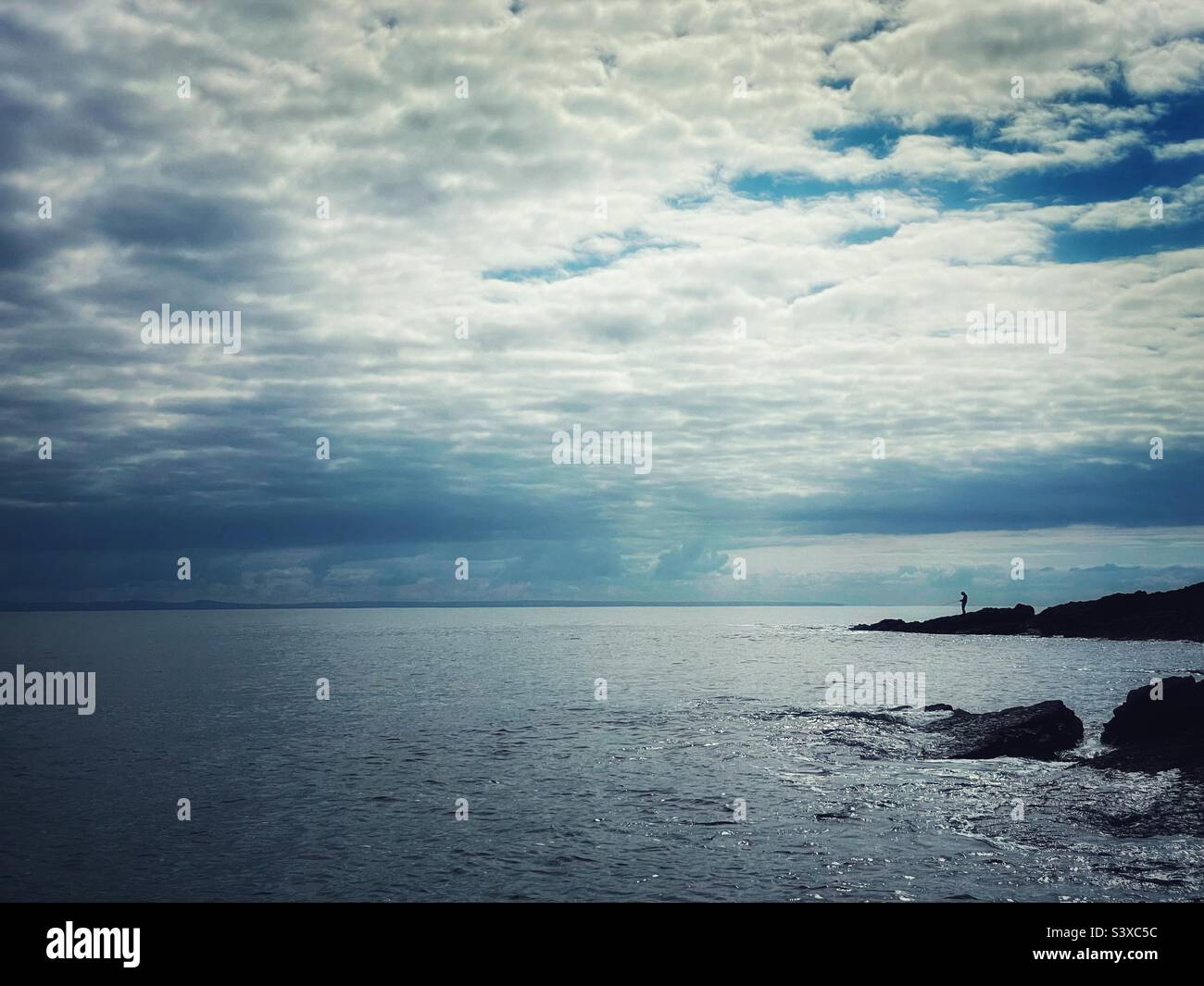Lone angler fishing from a rocky point on the coast of Wales, September. - Smartphone Captured Stock Image