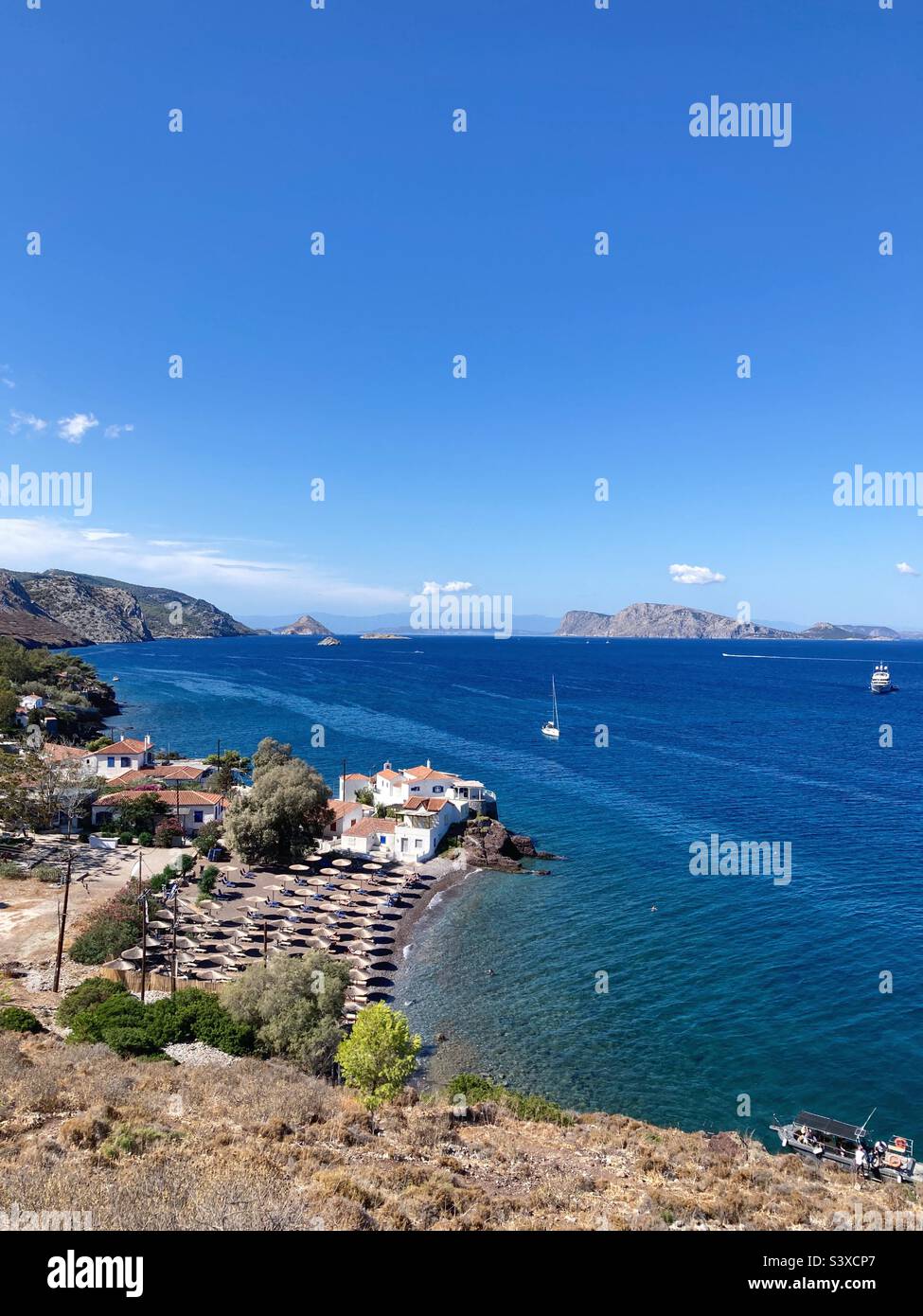 View of Vlychos Beach, Hydra, Greece, with chairs and parasols laid out and blue sky above - Smartphone Captured Stock Image
