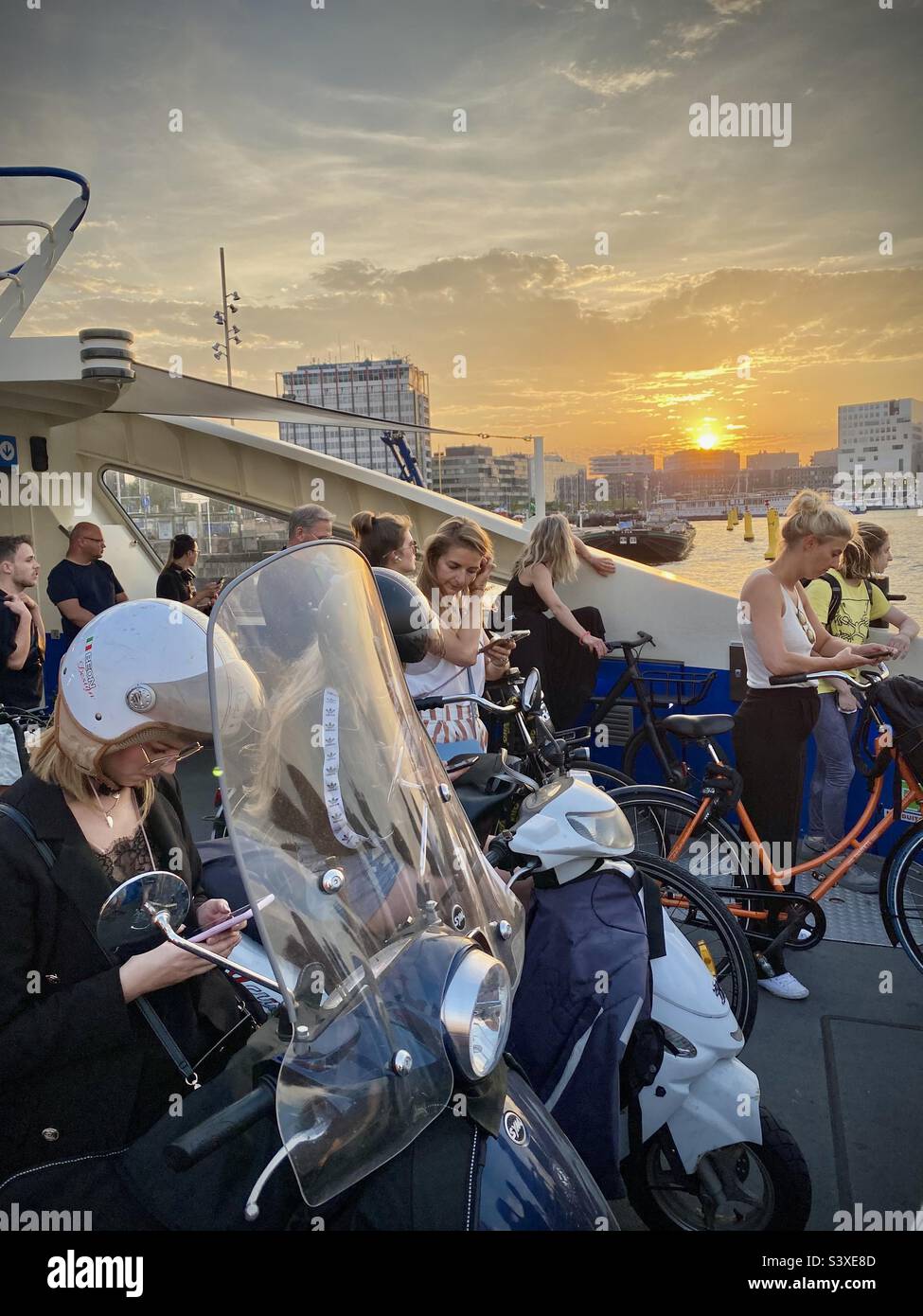 Amsterdam commuters on bicycles and mopeds leave the city centre on the free evening ferry and scroll their devices during a glorious sunset - Smartphone Captured Stock Image