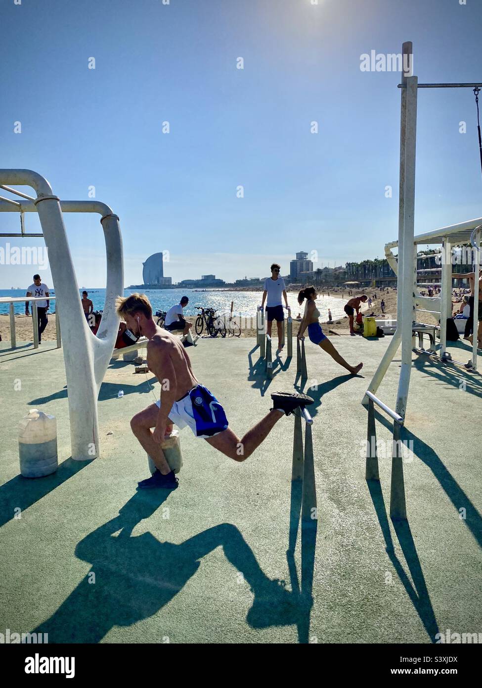 Athletes work out in public in the morning sun at the famous open air gym on the busy city beach of Barceloneta in Barcelona, Spain - Smartphone Captured Stock Image