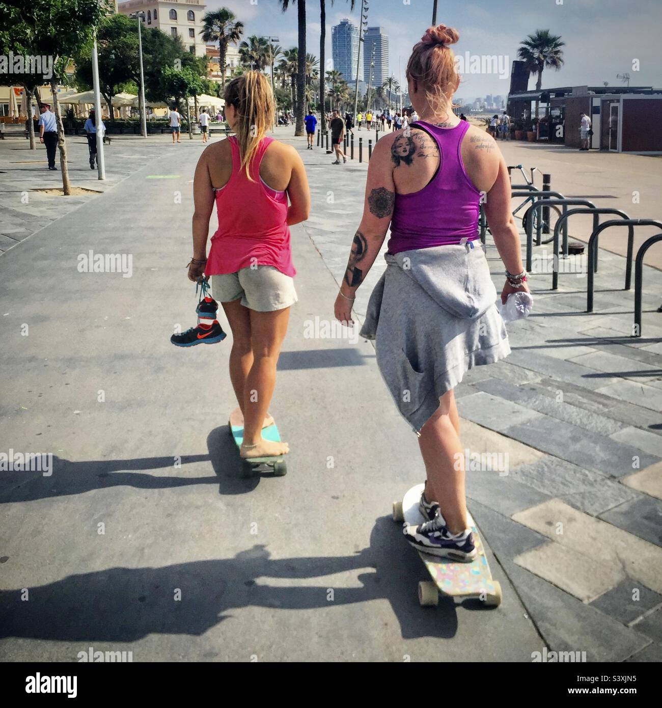 Two women in sport training gear skateboard together along the seafront of the busy city beach of Barceloneta in Barcelona, Spain Stock Photo