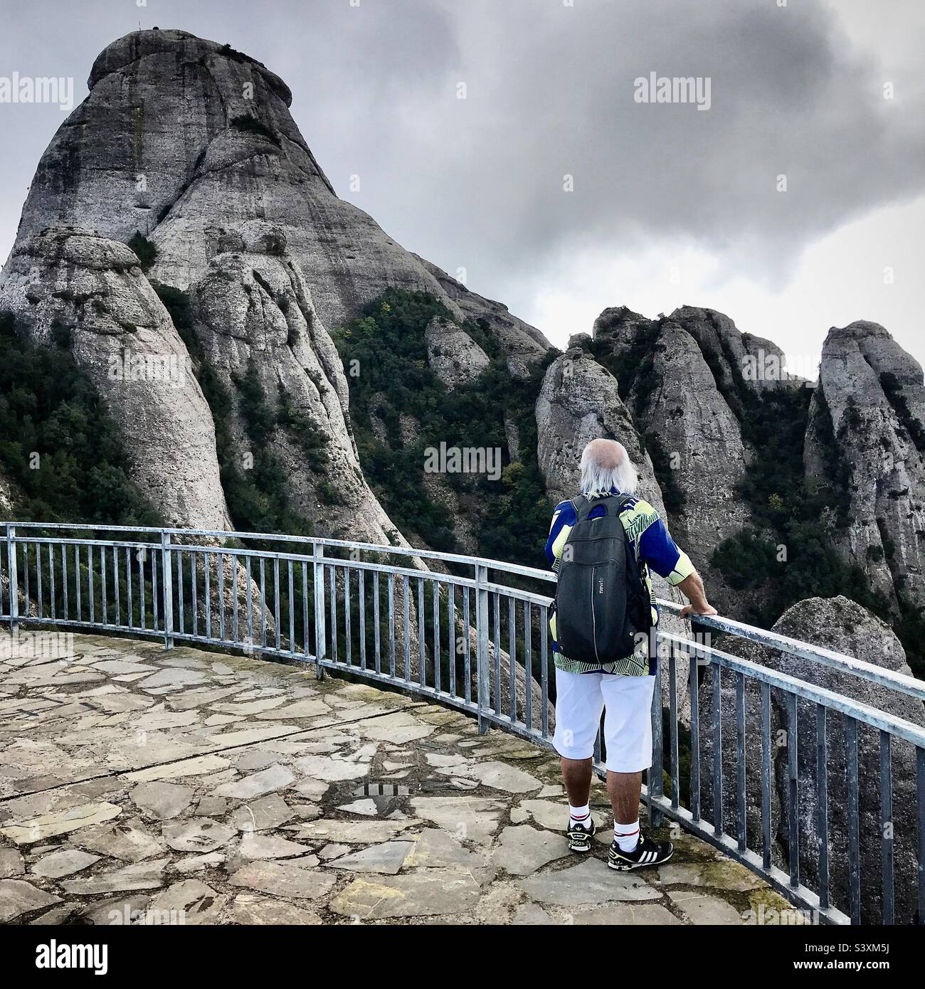 A mountain trekking tourist looks out from a mirador viewing platform on a public walking route through the Montserrat mountains near Barcelona in Catalonia in Spain - Smartphone Captured Stock Image