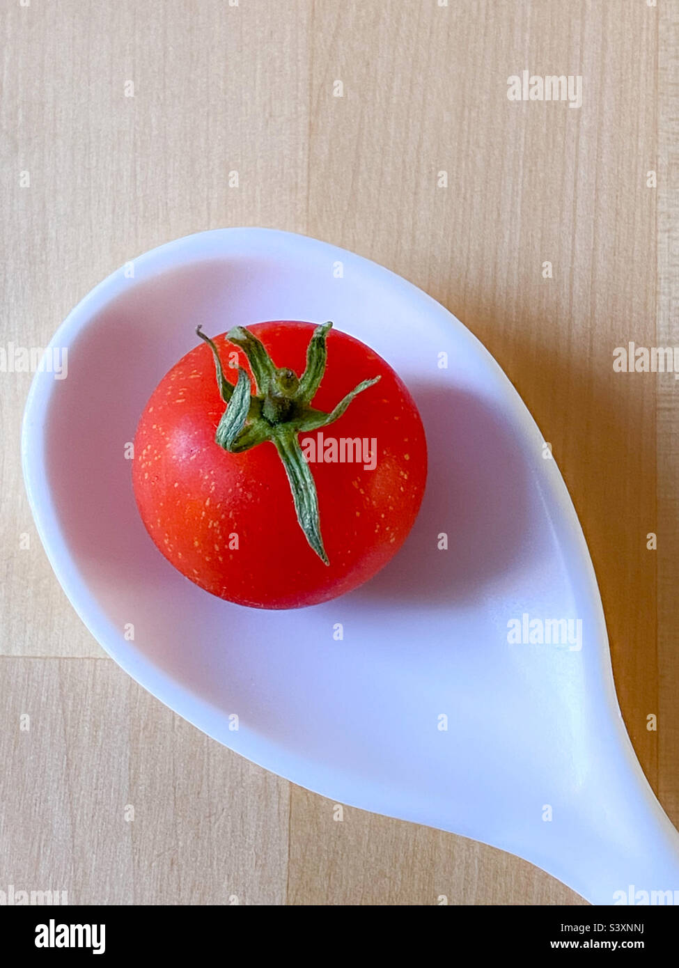 Still life image of a single cherry tomato in the bowl of a white spoon on a wooden table - Smartphone Captured Stock Image