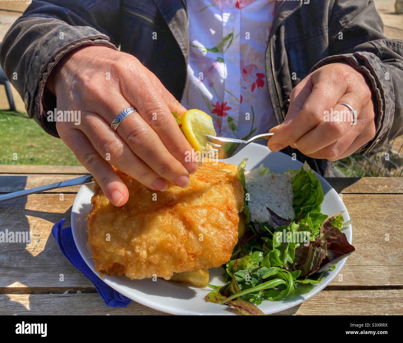 Fish and chips at an outdoor seaside cafe. Woman squeezing fresh lemon on to bartered fish. Midsection view with hands and plate. - Smartphone Captured Stock Image