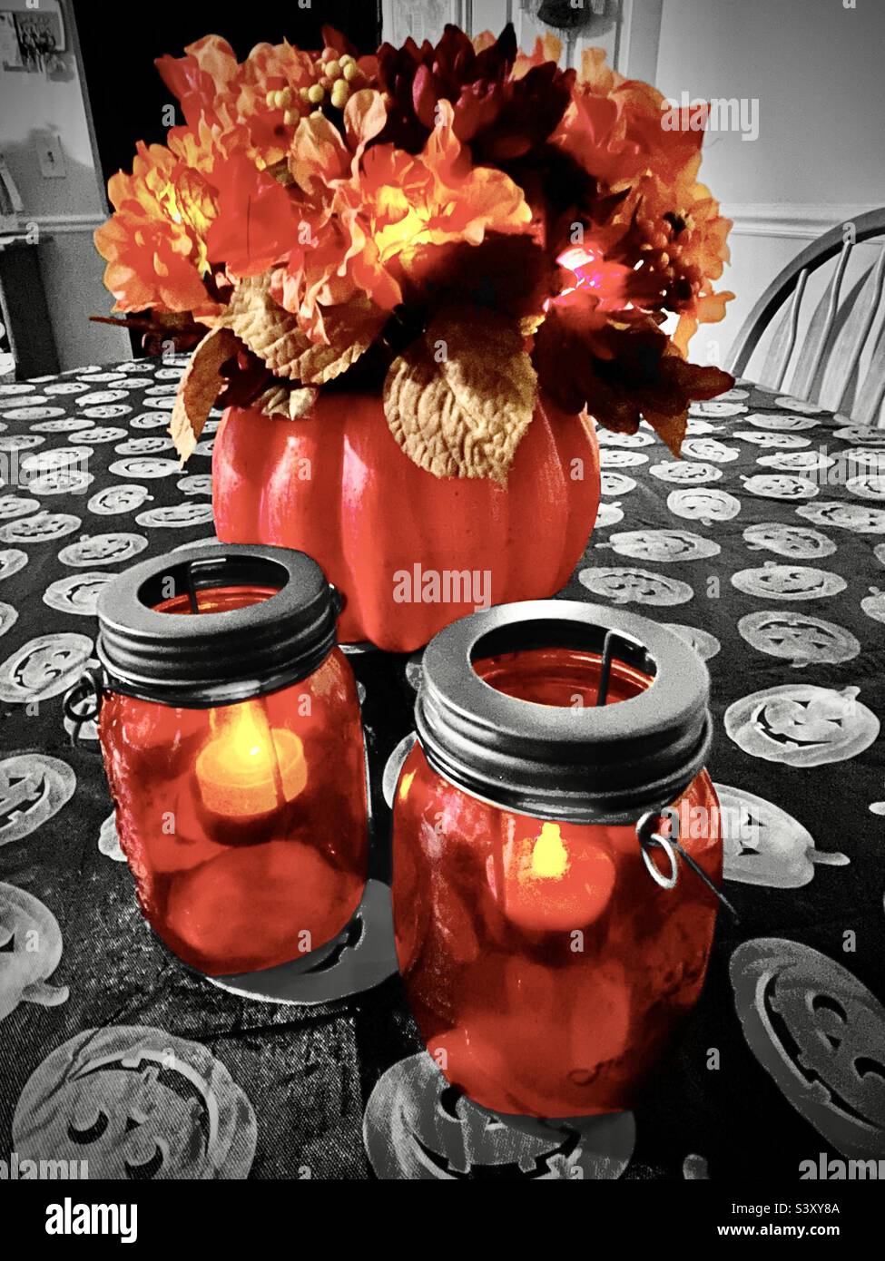 A tabletop set with Halloween and Autumn decor. The pumpkin floral arrangement and mason jar candles are battery lighted and color select, contrasting with the black & white Jack-o-lantern tablecloth. - Smartphone Captured Stock Image