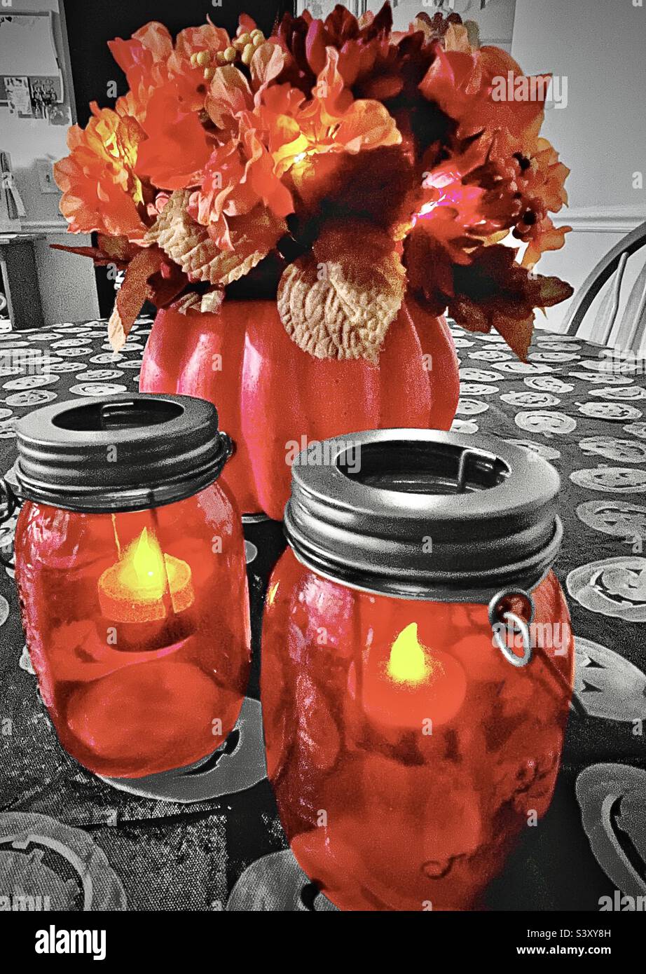 A tabletop set with Halloween and Autumn decor. The pumpkin floral arrangement and mason jar candles are battery lighted and color select, contrasting with the black & white Jack-o-lantern tablecloth. - Smartphone Captured Stock Image