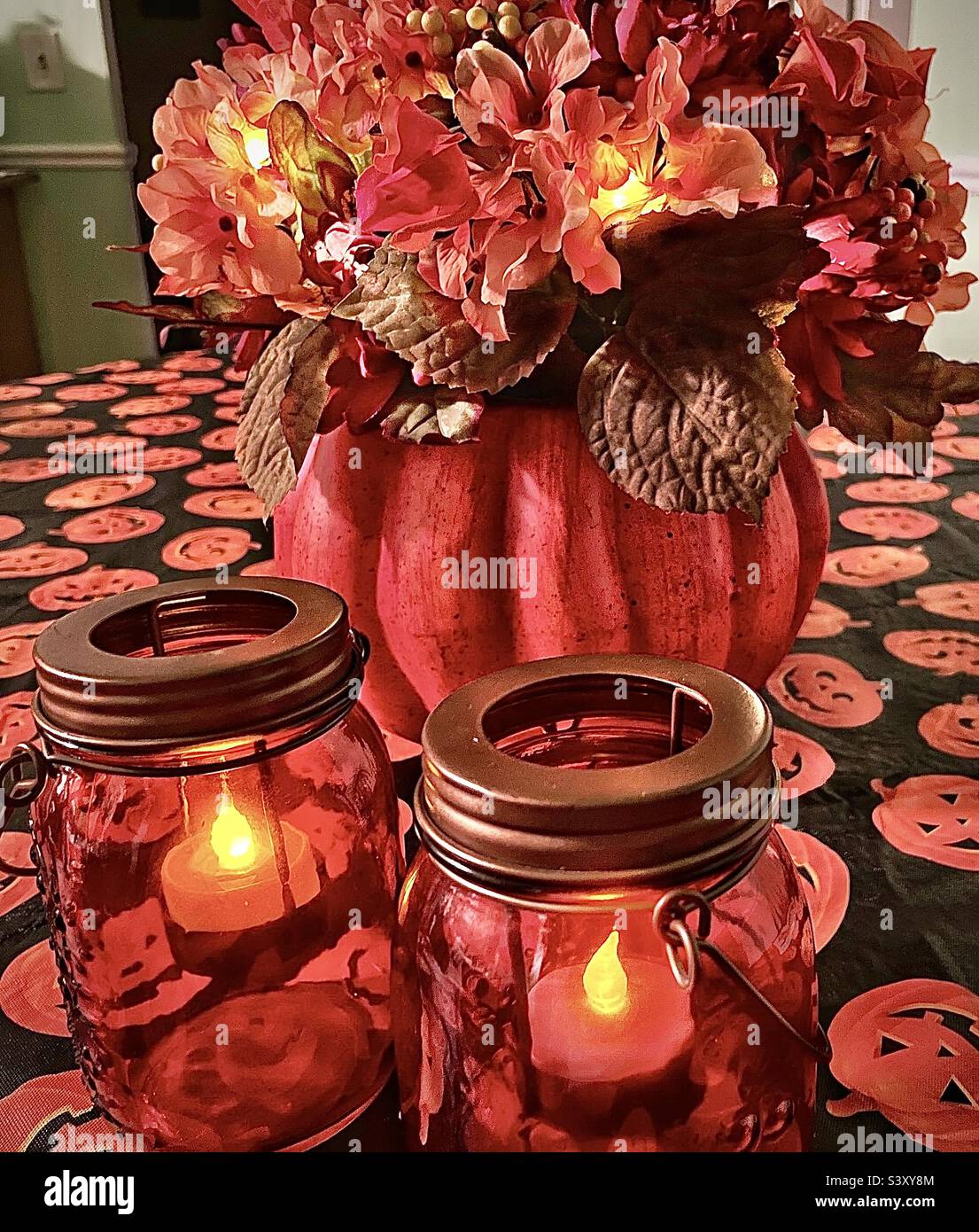 A tabletop set with Halloween and Autumn decor. The pumpkin floral arrangement and mason jar candles are battery lighted and sit atop a festive Jack-o-lantern tablecloth. - Smartphone Captured Stock Image