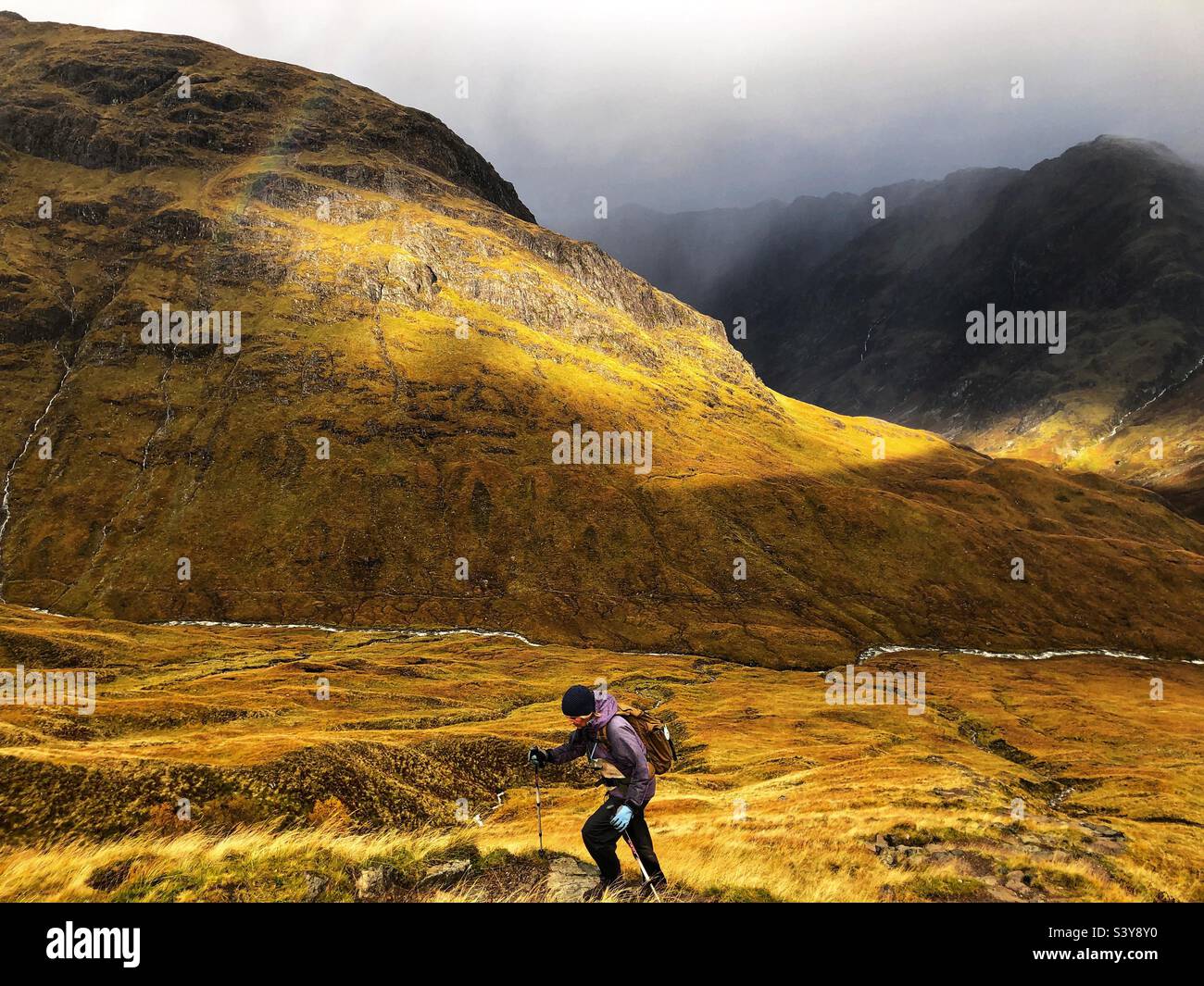 Female Walker with shafts of light as the clouds start to break on the path and slopes of Munro Buachaille Etive Beag, Glencoe, Scotland - Smartphone Captured Stock Image