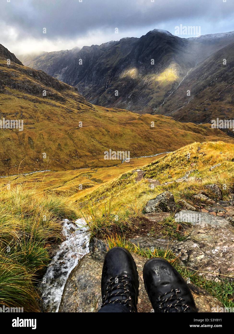 View of Walkers feet and boots with shafts of light as the clouds start to break on the path of Munro Buachaille Etive Beag, with a view towards the ridge of Aonach Eagach ridge, Glencoe, Scotland - Smartphone Captured Stock Image