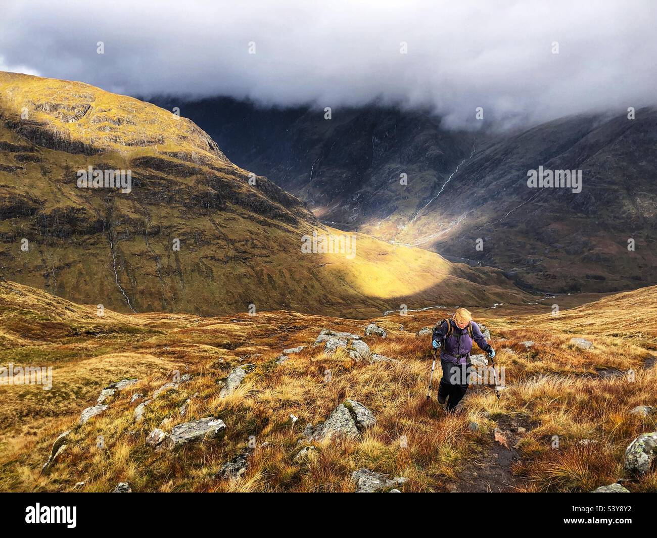 Female Walker with shafts of light as the clouds start to break on the path and slopes of Munro Buachaille Etive Beag, Glencoe, Scotland - Smartphone Captured Stock Image