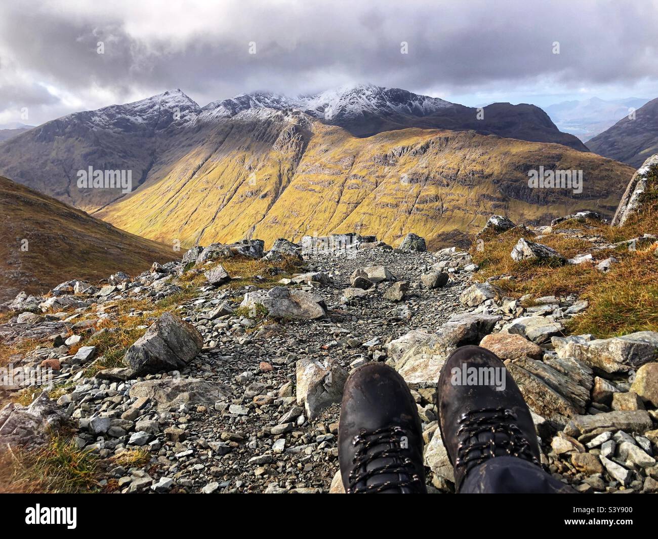 View of Walkers feet and leather boots on the path and slopes of Munro Buachaille Etive Beag, with a view of snow capped peaks of Bidean nam Bian, Glencoe, Scotland - Smartphone Captured Stock Image