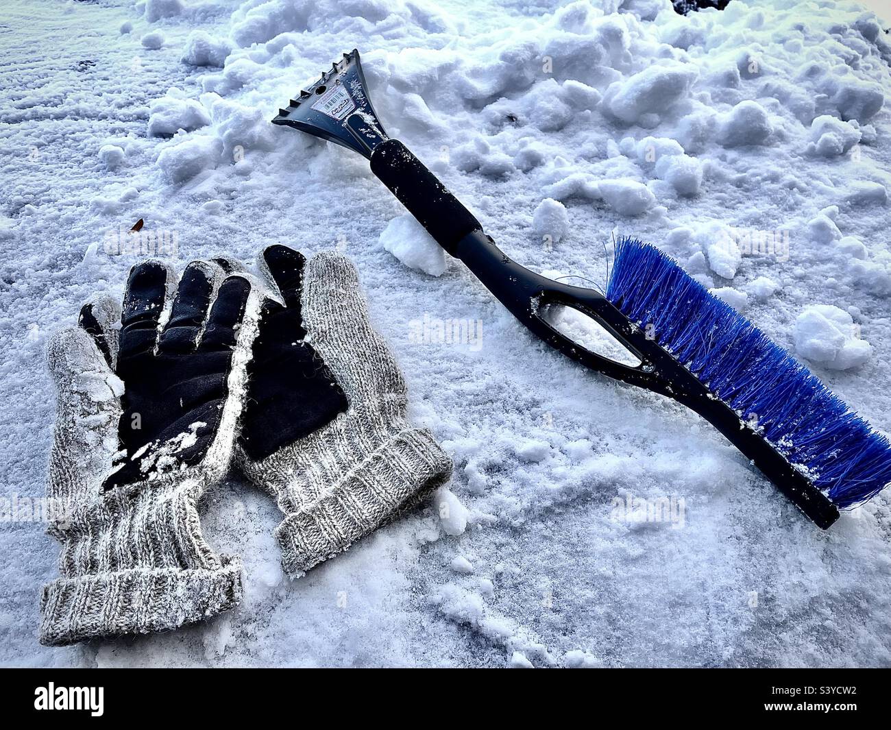 A pair of wool gloves and a snow/ice tool, one end an ice scraper, the other end a snow brush. A wintry still life set upon a snow covered car hood after a Utah, USA snowstorm. Stock Photo