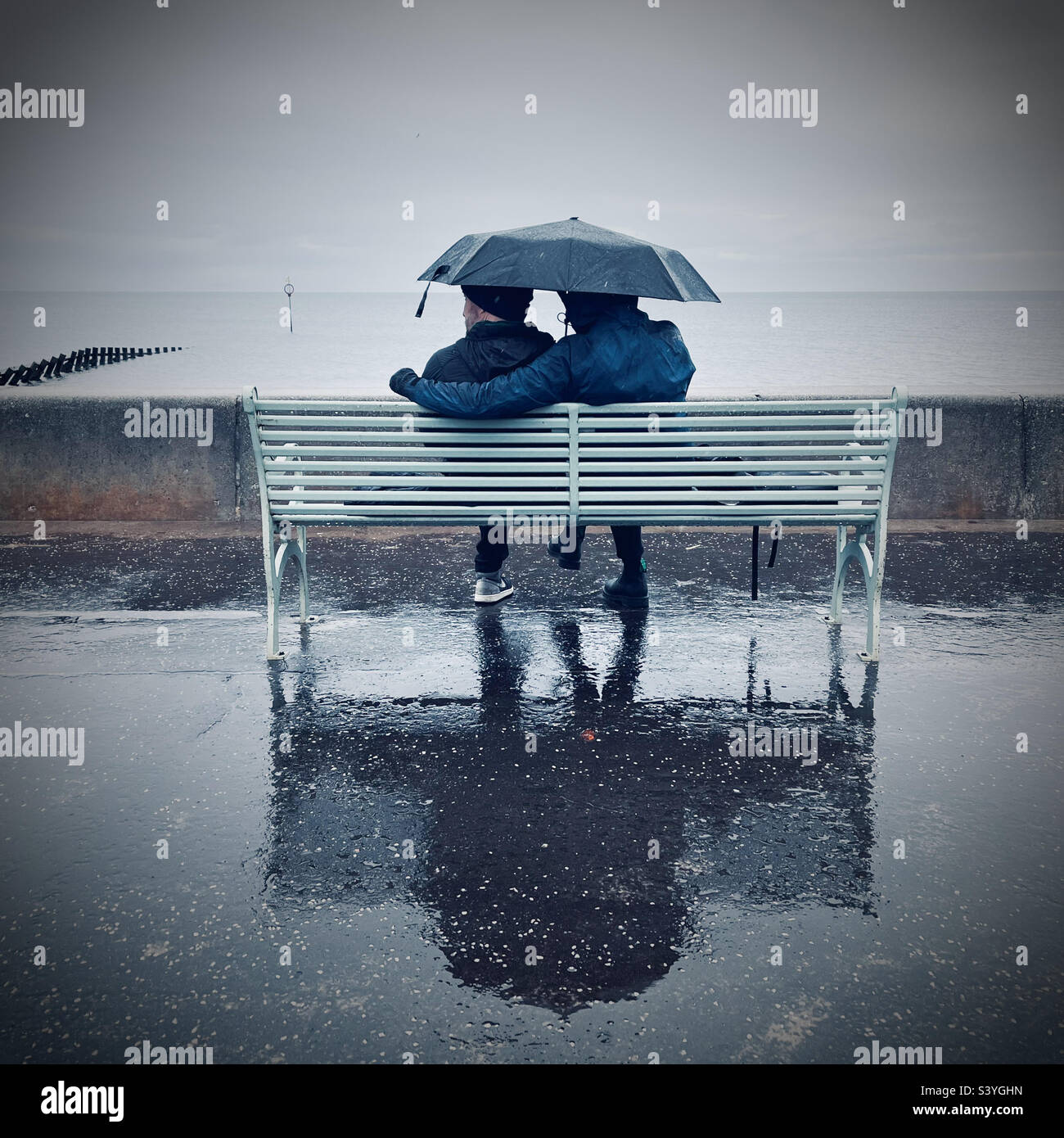 Couple sitting on bench in the rain looking out to sea - Smartphone Captured Stock Image