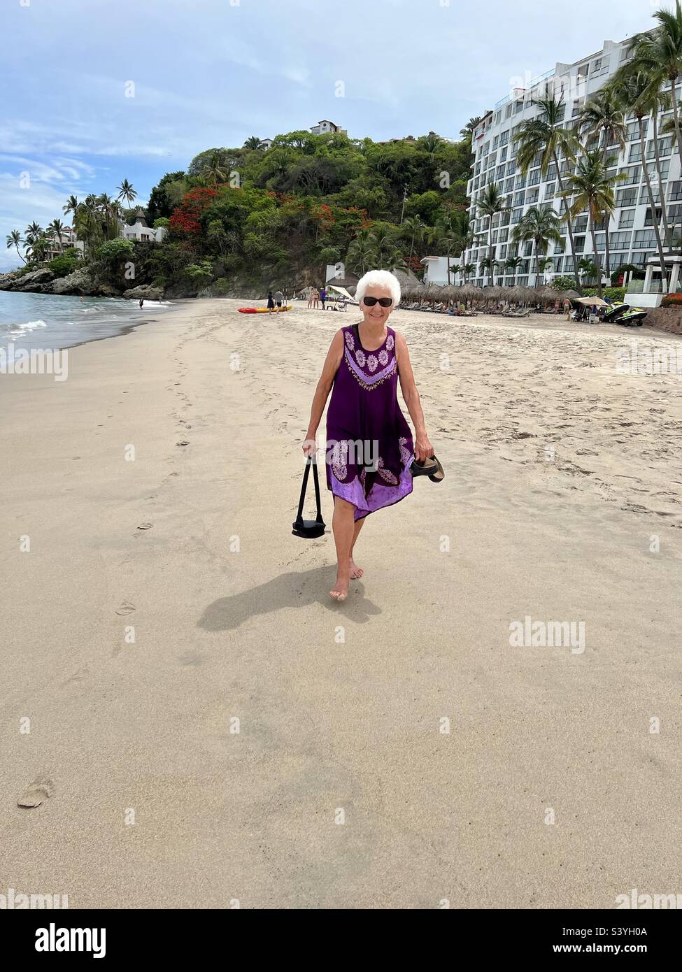 A senior Woman with white hair or silver hair walking on a beach in a purple sundress Stock Photo