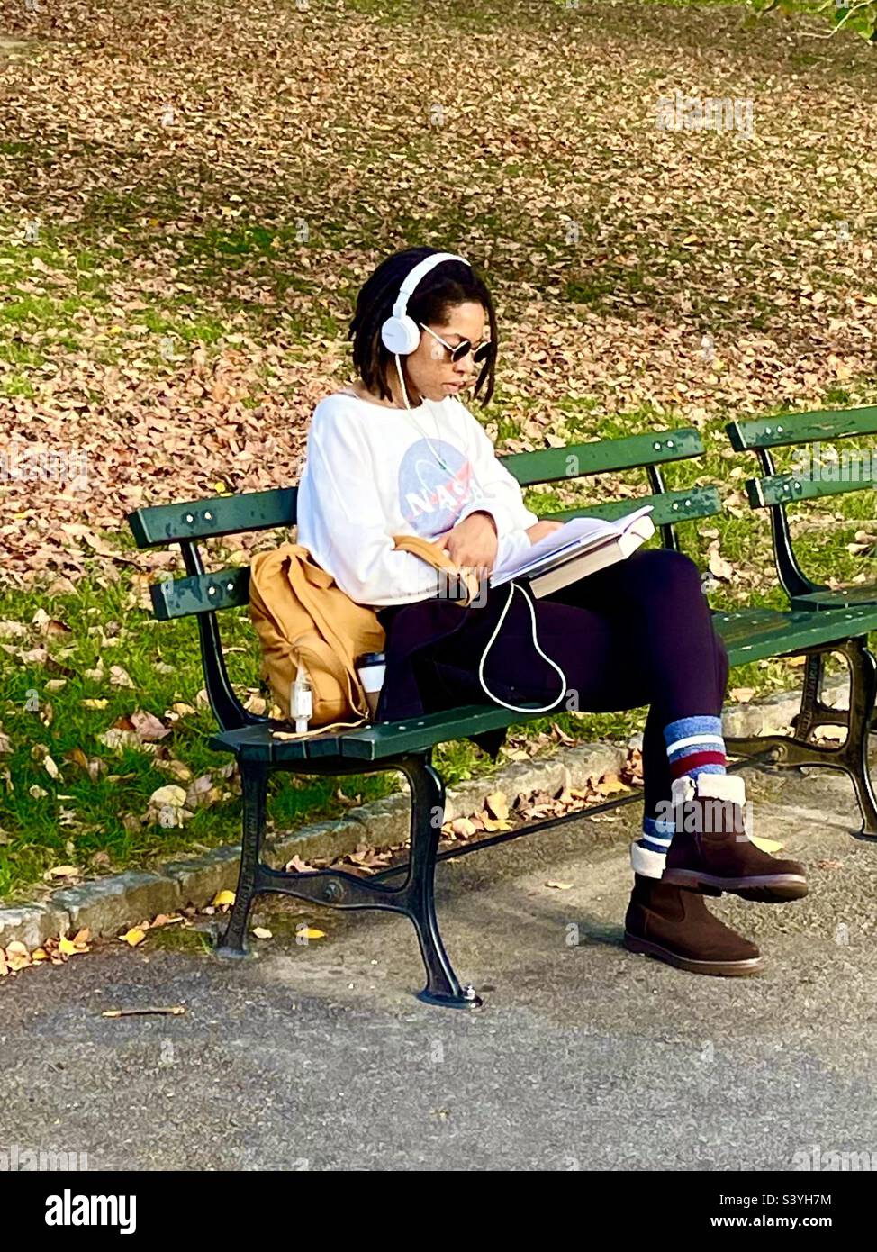 A young woman sitting on a bench reading a book with earphones phones - Smartphone Captured Stock Image