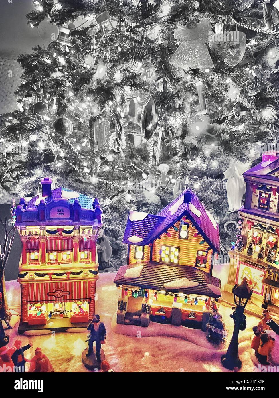 A lighted vintage village is part of a home’s Christmas decor. Old-fashioned town buildings, and town residents, glow colorfully, while the home’s Christmas tree is desaturated in the background. - Smartphone Captured Stock Image