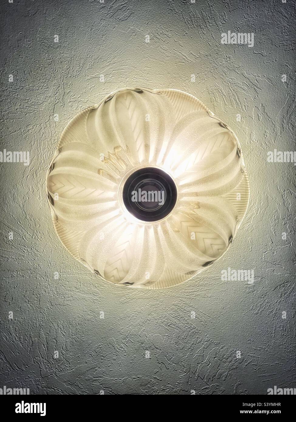 Shot directly from below, this decorative glass light fixture emits a soft, warm glow and creates a central symmetrical design while also bringing out the ceiling texture. - Smartphone Captured Stock Image