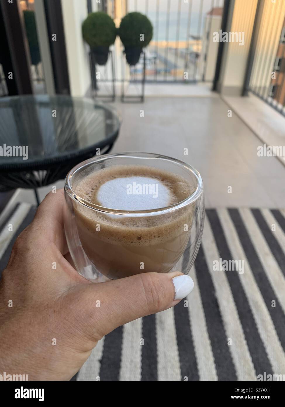 Woman holding a cup of cappuccino coffee on a balcony overlooking the sea - Smartphone Captured Stock Image