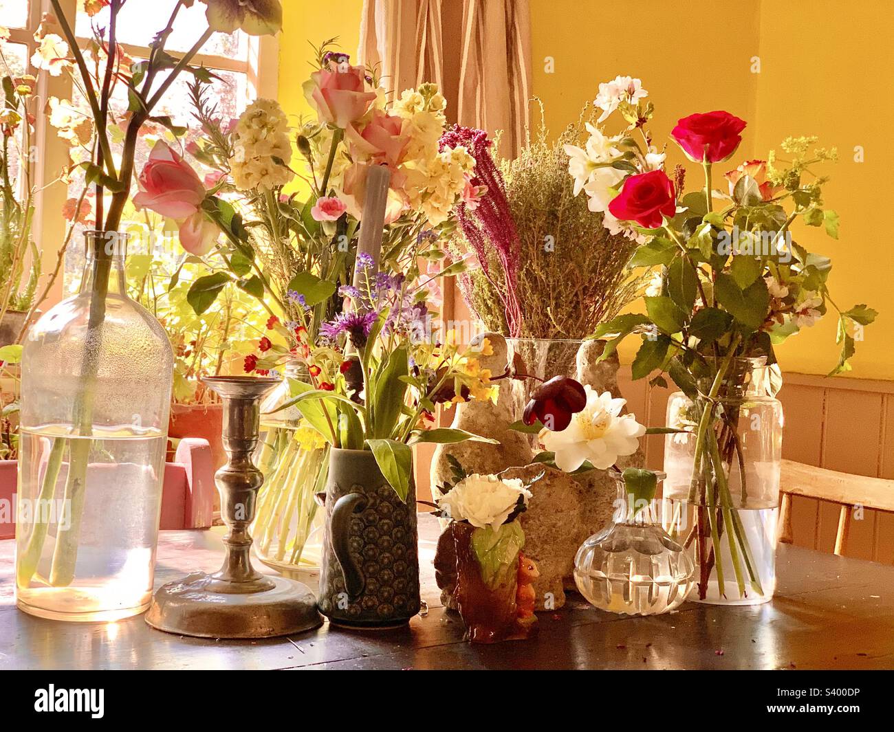 Beautiful cottage kitchen tablescape with cut flowers in a variety of glass jars and ceramic vases with decorative candlestick in bright yellow countryside kitchen in spring, England Stock Photo