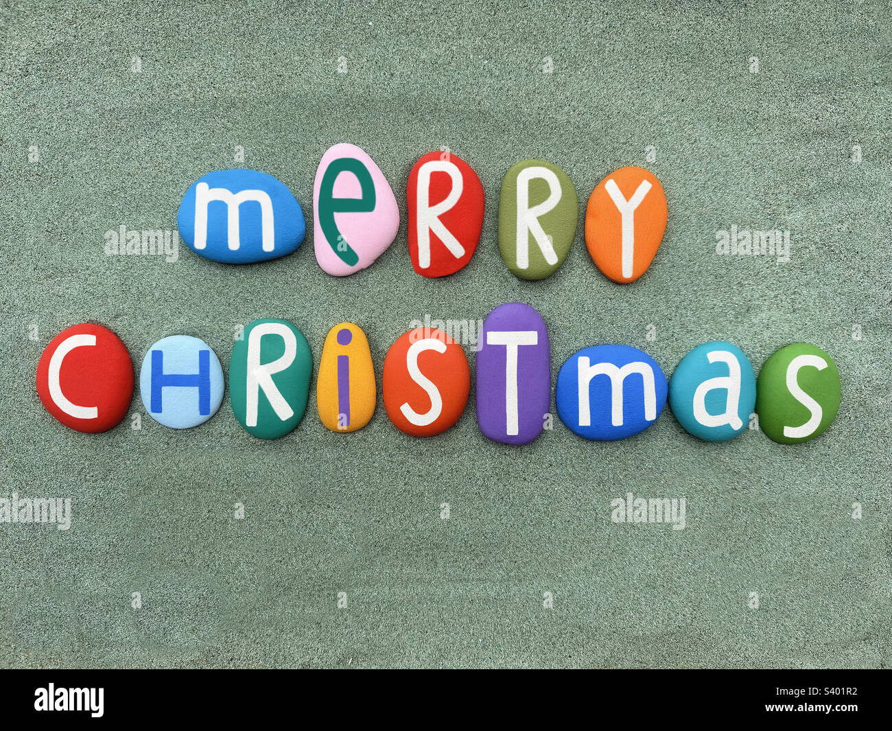 Merry Christmas message composed with multi colored hand painted stone letters over green sand - Smartphone Captured Stock Image