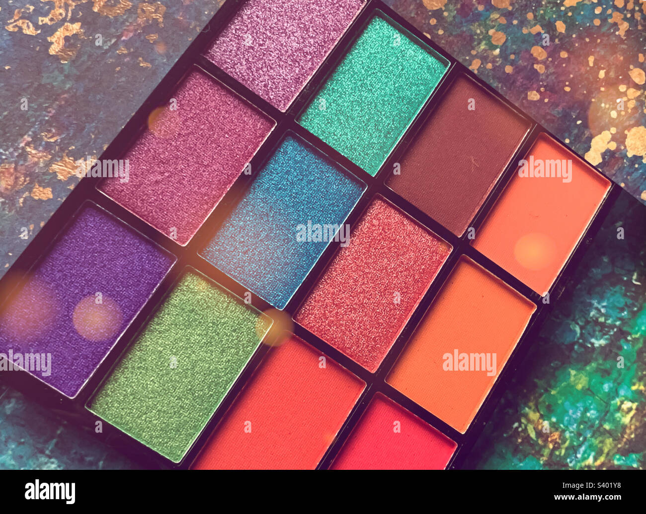 A vibrant palette of colourful eyeshadows and lipgloss against a patterned background, with added bokeh - Smartphone Captured Stock Image