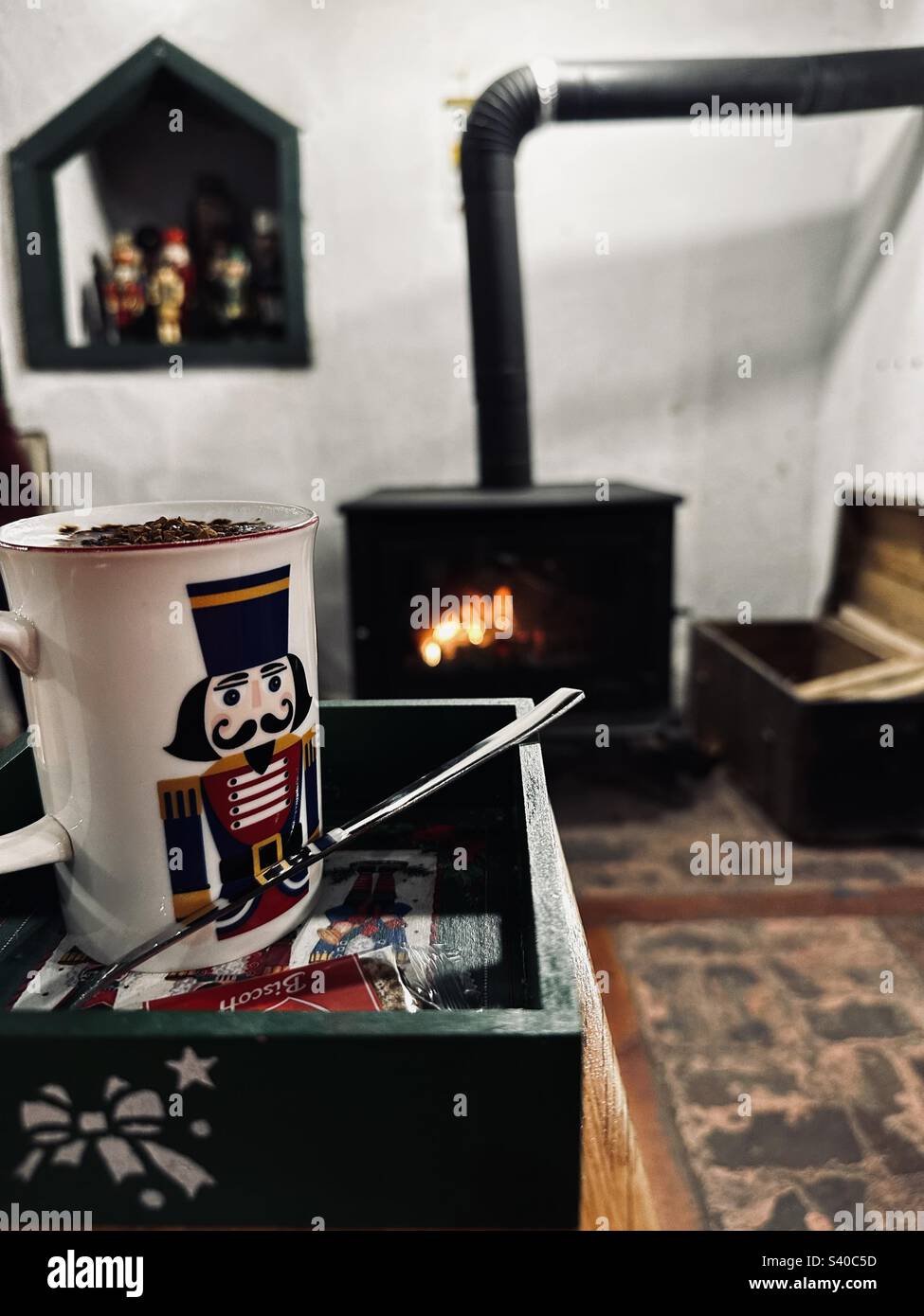 Coffee in a mug having tin soldier print served together with Biscoff. On the background, there is a heated stove and a wooden box storing firewoods and tin soldier statues in the hole on the wall - Smartphone Captured Stock Image