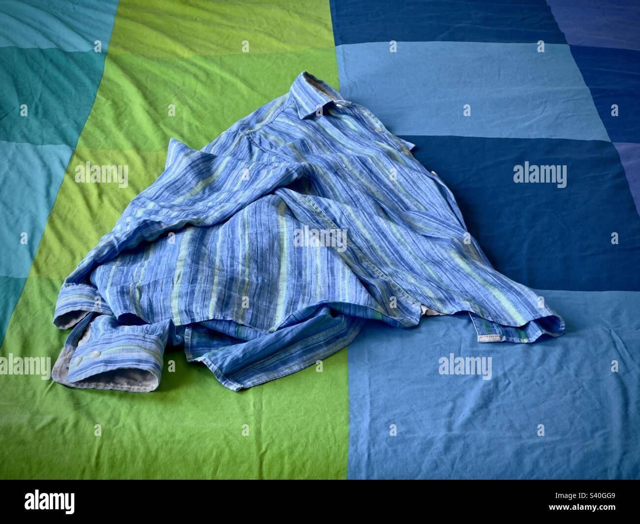 Wide shot. Blue, green and indigo linen shirt unfolded, dropped on a blue, green and indigo bed cover. Whole shirt is visible - Smartphone Captured Stock Image
