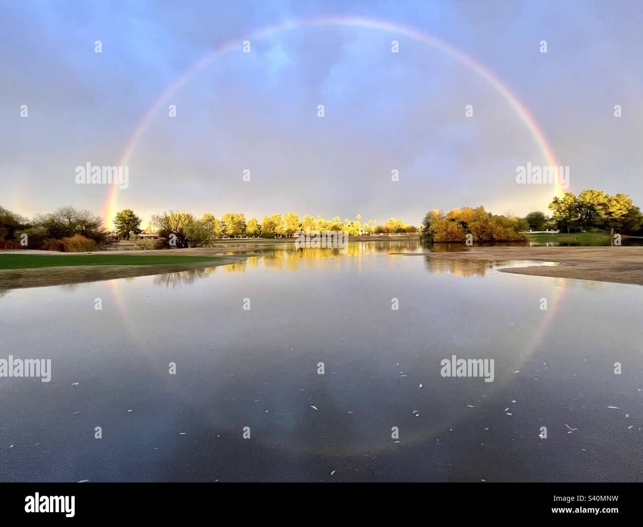 Head on, 360 degree rainbow, circling infinity perspective. Rainbow reflection in pond, tree reflections, stormy blue sky, golden sunset hues Stock Photo