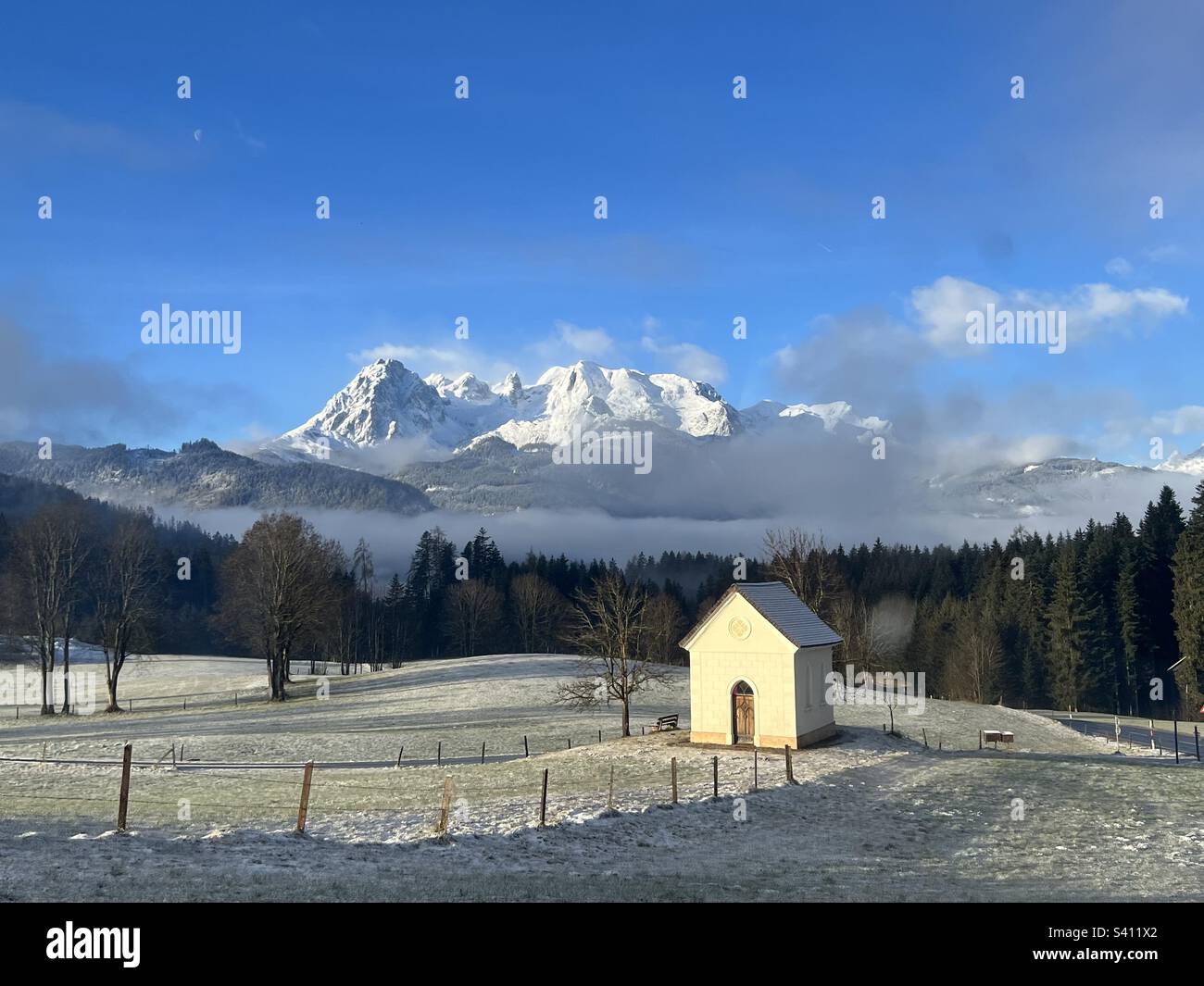 Image of a small church in Austria surrounded by fog and twigs on the meadow;  in the background forest and a beautiful mountain landscape Stock Photo