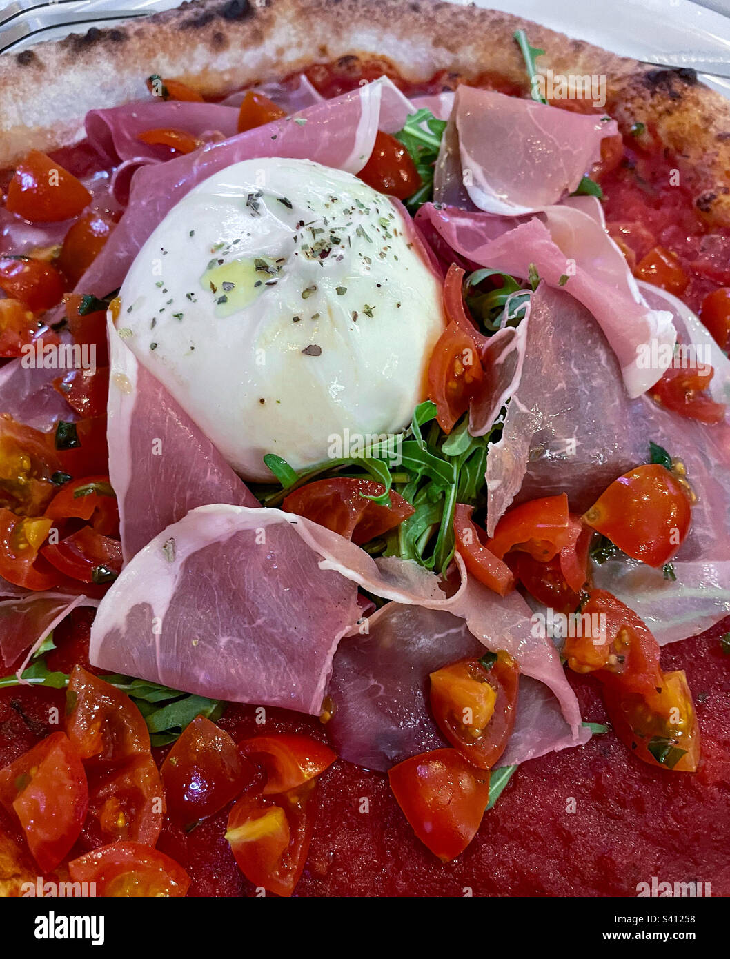 Close-up of Italian pizza with buffalo mozzarella, chopped tomatoes, prosciutto ham, arugula, sprinkled with oregano and olive oil on a thin crust with thick tomato sauce, photographed from overhead Stock Photo