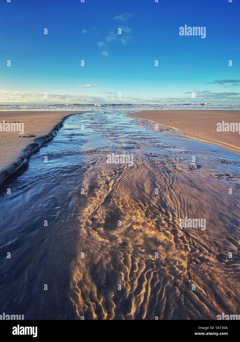 A photograph taken low looking down a stream on a Sandy beach flowing towards the ocean. Ripples and reflections of fresh water on a beauty winter morning - Smartphone Captured Stock Image