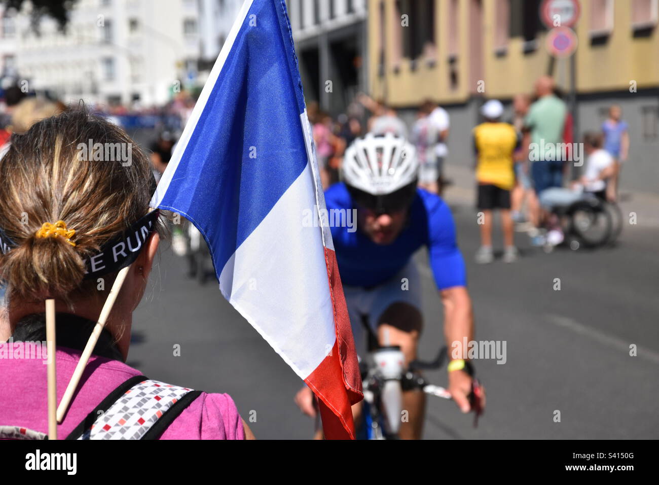 A female spectator from rear view with a French flag looking towards an triathlon athlete on his bike. Stock Photo