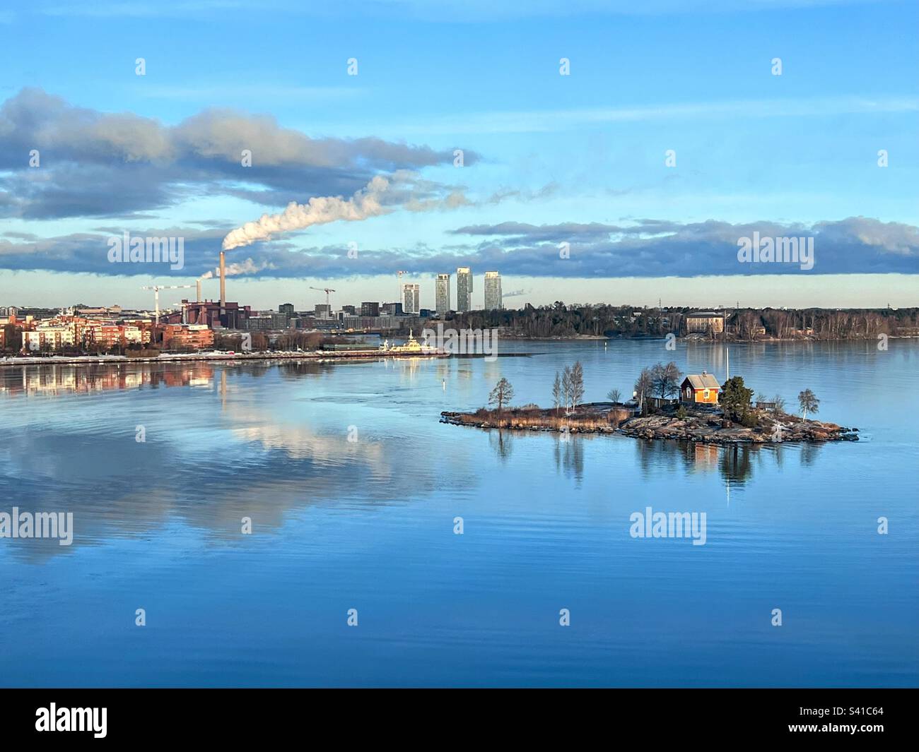 Tranquil scene with Helsinki coastline cityscape with smoking pipe, little island with trees and small wooden cottage and clouds reflecting in water - Smartphone Captured Stock Image