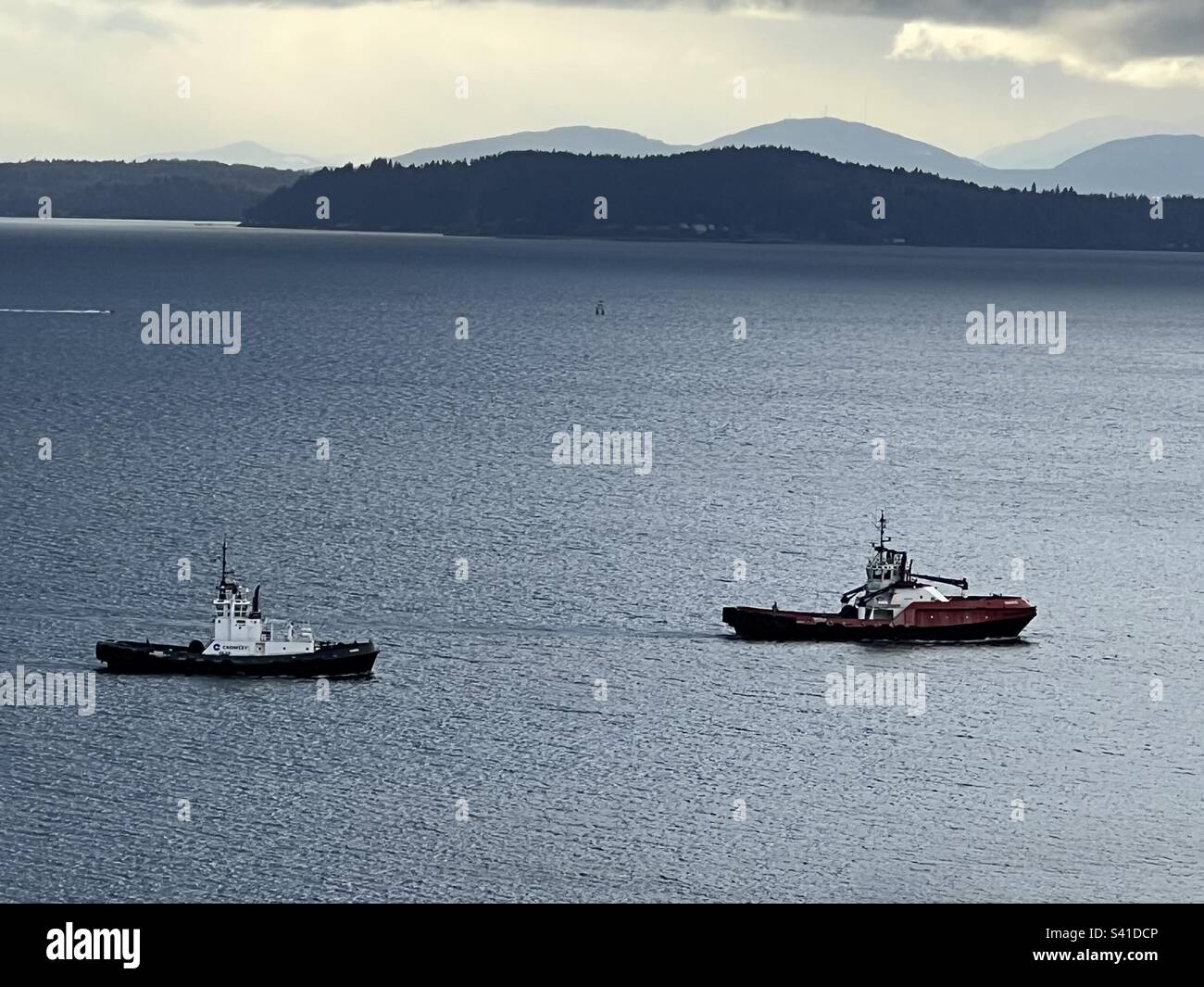 Two tugboats waiting for container ship to arrive so they can help dock to unload at Seattle harbor, Elliot Bay, Puget Sound, WA State, PNW, USA. Olympic Mountains in background across Salish Sea. Stock Photo