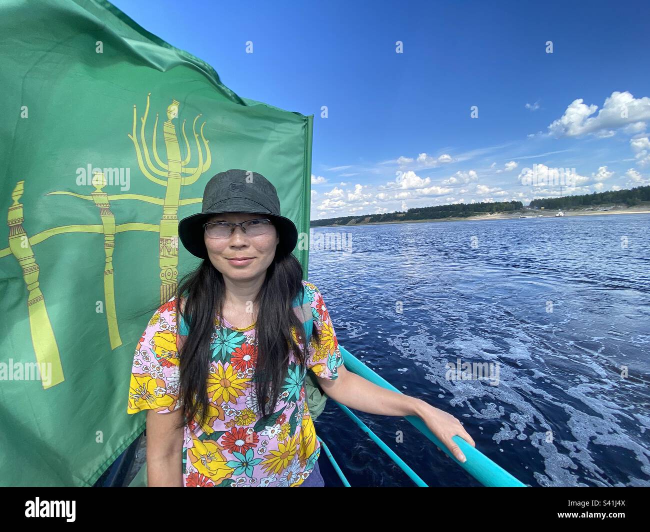 An Asian girl Yakut stands in glasses and a hat smiling on board a ferry going down the river on the background of the flag. Stock Photo