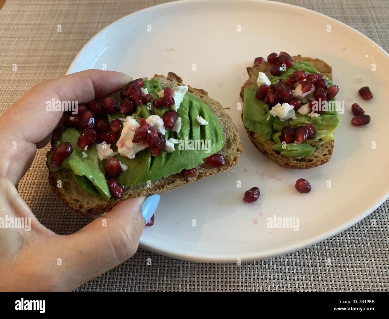 Woman holding a slice of toasted sourdough bread topped with avocado, feta and pomegranate - Smartphone Captured Stock Image
