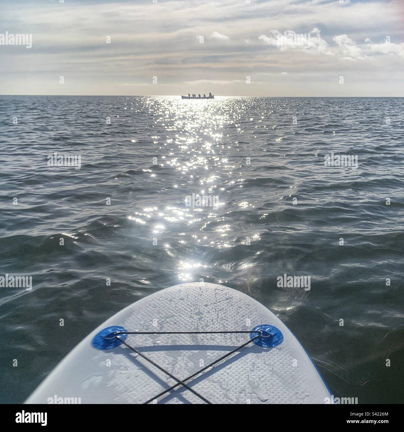 View of a pilot gig rowing boat highlighted by the sun from a stand up paddle board in the sea at Sidmouth Stock Photo