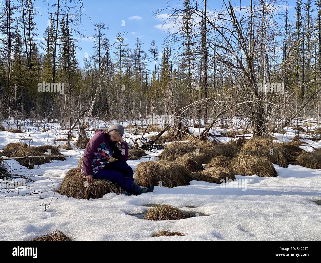 An Asian girl Yakut is sitting, bowing her head and smiling, on a tussock overgrown with grass in the spring in the forest of Yakutia Stock Photo