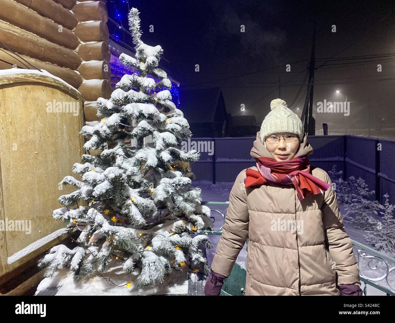 A frozen Yakut Asian girl with glasses stands in winter in the cold at a Christmas tree with snow in the village of Yakutia. Stock Photo